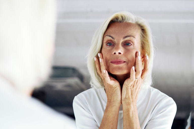 gettyimages-2199411062-1748872487 Close up of older 65s woman stand in front of mirror, gently touch her face with hands, examining skin, assessing results of skincare routine. Hydration, anti-ageing regimen, aging gracefully concept
