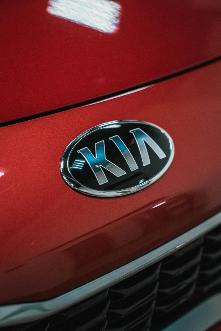 kkk-1780316641 Detailed view of KIA emblem on sleek red car hood, showcasing modern automotive design.