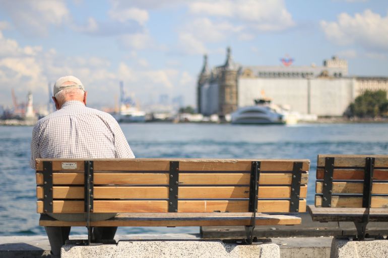 msnwn-1781627220 An elderly man sits alone on a bench by the waterfront, enjoying a peaceful day.