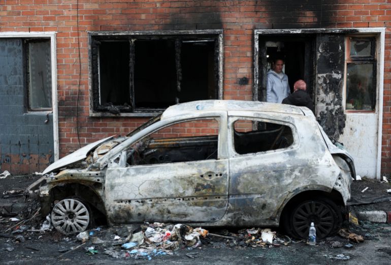 reuters_6a2a39bb-1781152187 A woman speaks to a person from the doorway of her burnt house, as a destroyed car stands on the street, in the aftermath of anti-immigrant protests following a knife attack on June 8, which left a man seriously injured and prompted police to declare a critical incident, in Belfast, Northern Ireland, June 10, 2026. REUTERS/Isabel Infantes