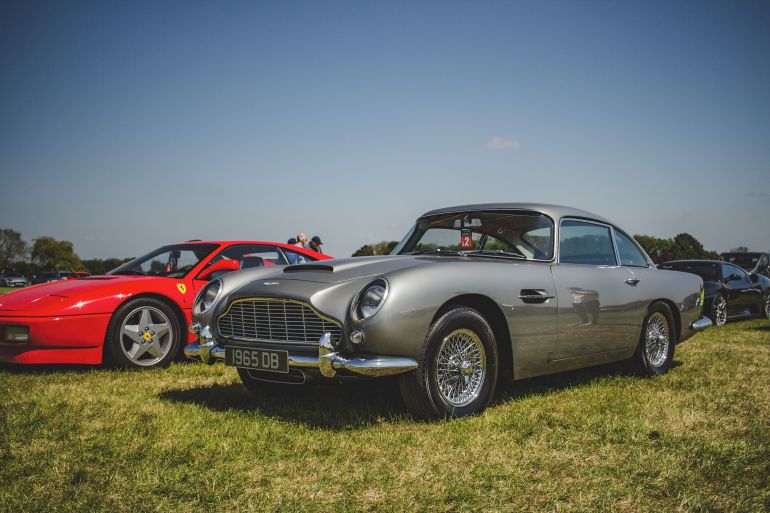 shhyrt-1780730431 Vintage Aston Martin and red Ferrari parked on green grass under bright daylight.
