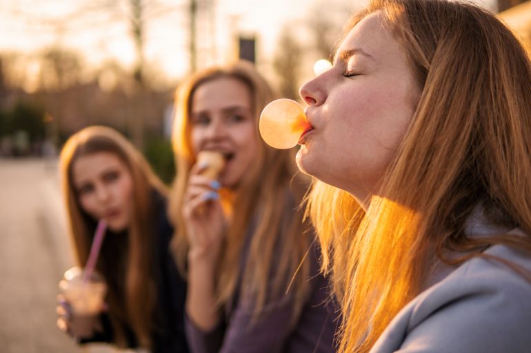 Three girls friends having fun on evening broadway. Eating ice cream, drinking ice fresh milk shake and blowing chewing bubble gum
