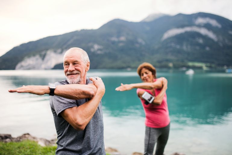 shutterstock_1537647920-1780304027 Senior pensioner couple with smartphone by lake in nature, doing exercise.