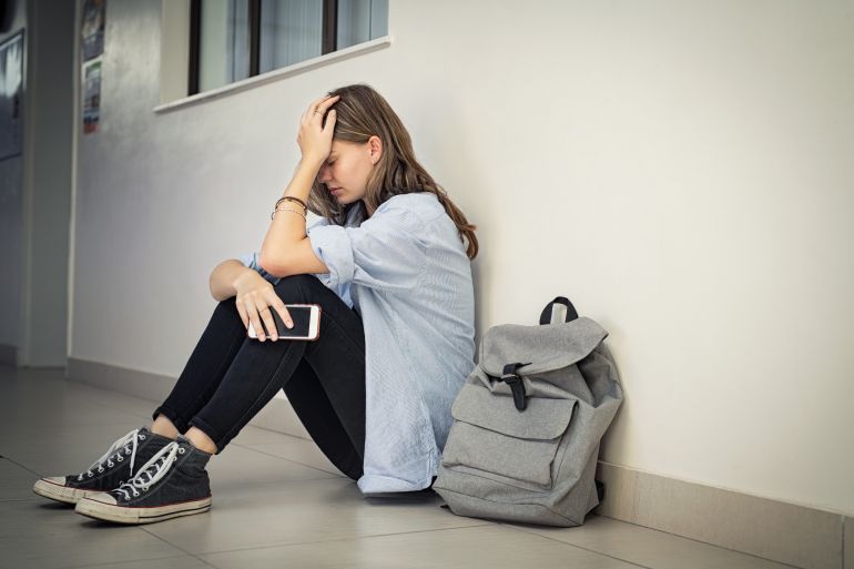 shutterstock_1833171139-1743021017 Upset and depressed girl holding smartphone sitting on college campus floor holding head. University sad student suffering from depression sitting on floor at high school. Lonely bullied teen.