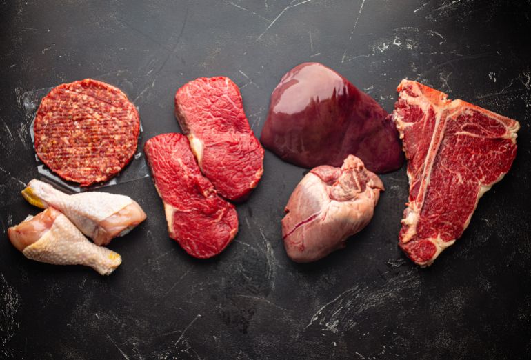 shutterstock_1958573278-1781087768 Selection of assorted raw meat food for zero carb carnivore diet: uncooked beef steak, ground meat patty, heart, liver and chicken legs on black stone background from above