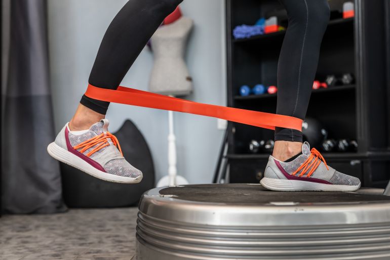 shutterstock_2169733539-1780911091 Feet of unknown woman and rubber elastic resistance band while standing on power plate vibration platform machine during training workout exercise copy space