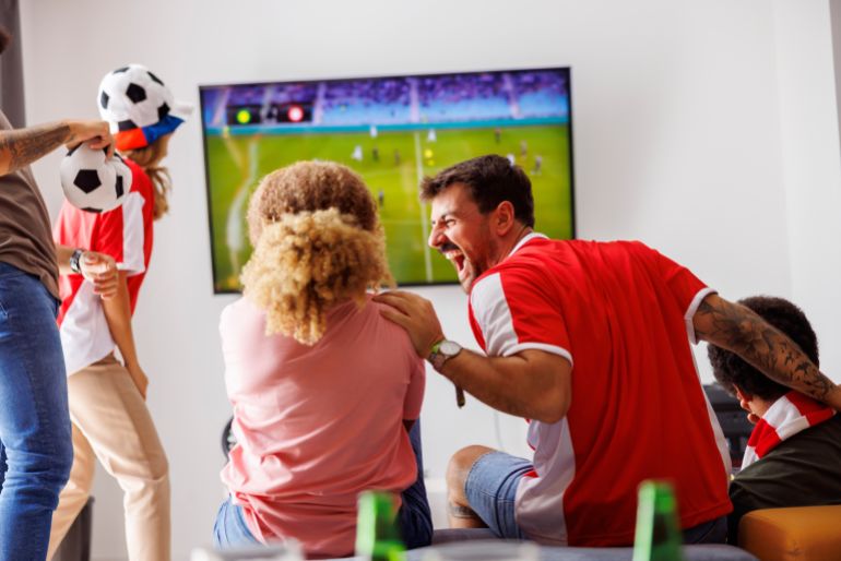 Group of young friends having fun watching football match on TV, drinking beer and cheering; football fans watching game at home celebrating after their team scoring a goal
