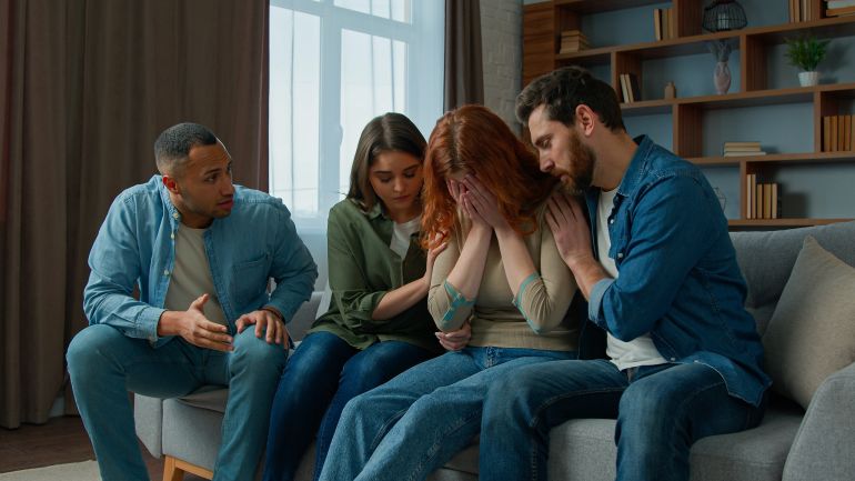 shutterstock_2295533667-1776846734 Sad desperate girl talk share problem with friends in group psychological therapy at home diverse ethnic women men help upset girl. Caring multiracial teammates listen complaints support give advice
