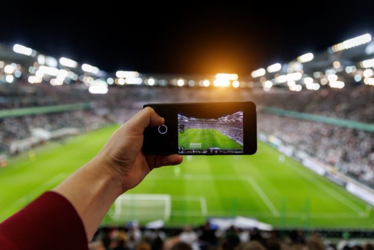 shutterstock_2369769949-1780913903 Fan hand with smartphone photographing football match. Using mobile phone camera at the stadium