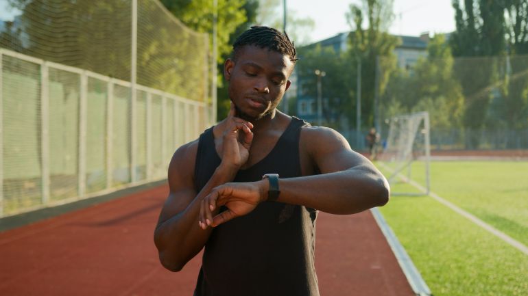 shutterstock_2387041681-1780303493 African American man runner jogger athlete muscular sportsman training outdoors athletic ethnic guy using smart band checking neck pulse heart rate result on smartwatch before running on sport stadium