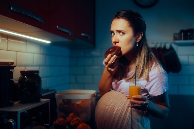 shutterstock_2401944293-1738153549 Pregnant Woman Eating Chocolate Doughnuts Drinking Orange Juice. Funny hungry mother to be snacking during nighttime