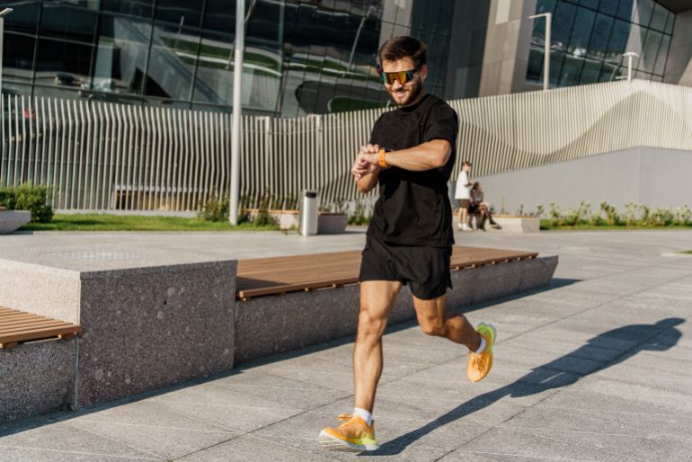 shutterstock_2495911501-1780304313 A man running in a modern urban area, checking his smartwatch, wearing athletic gear and sunglasses.