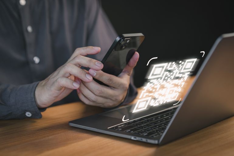 shutterstock_2544622157-1780500058 Person uses a smartphone to scan a QR code displayed on a laptop screen, symbolizing digital transactions, authentication, and modern contactless technology for secure access.