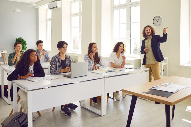 Male young friendly teacher at the lesson in high school. Students and classmates sitting at the desks listening their lecturer and working in modern classroom with laptop. Education concept.; Shutterstock ID 2592149727; purchase_order: aj; job: ; client: ; other: