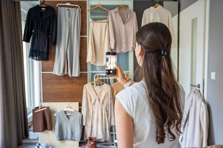 shutterstock_2596611295-1777638661 A young woman photographs clothing hanging on a wardrobe in bedroom using her smartphone. Online resale and second-hand fashion thrive through mobile apps, making sustainable shopping more accessible.