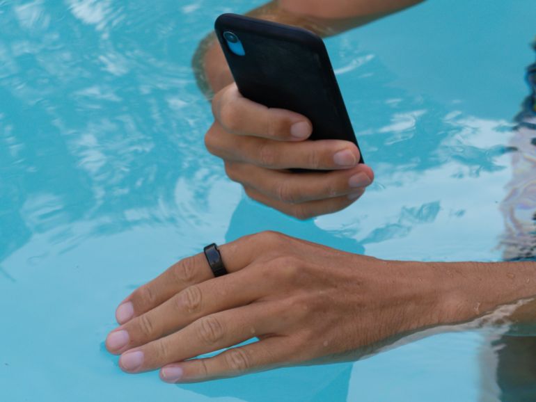 shutterstock_2635371677-1781004690 Hands in pool connect black smart ring health tracker to smartphone app.