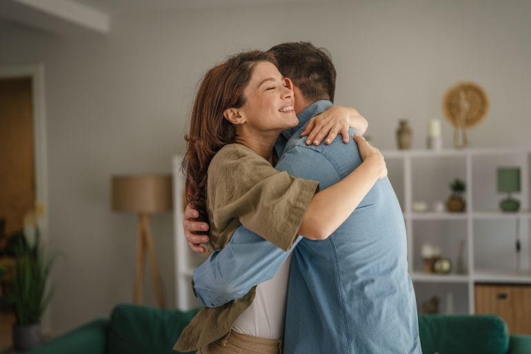 shutterstock_2737872537-1778238856 Happy couple embracing in a warm, loving hug at home, sharing a moment of affection, connection, and togetherness in their living room, representing comfort and reunion