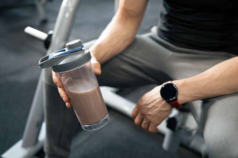 Athletic mature man holding chocolate protein shake after workout in modern fitness club.