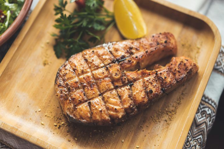 smk-1779694813 Close-up of seasoned grilled salmon steak with lemon and herbs on a wooden platter.