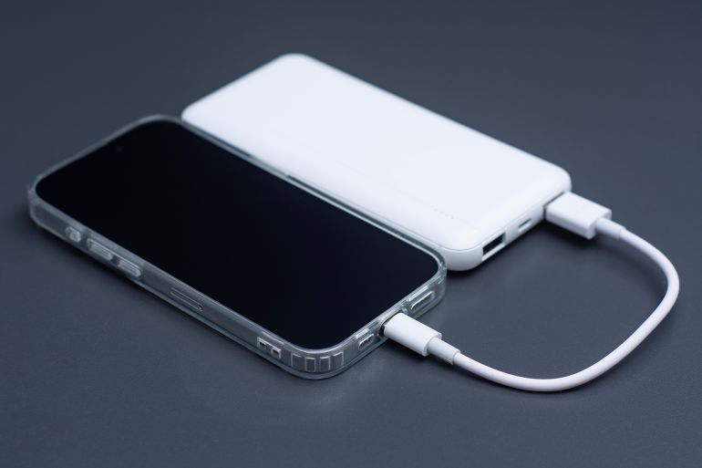 3-1783146243 Mobile phone connected to white external power bank with usb cable on dark background. Portable backup battery charging smartphone screen mockup for travel, technology or emergency energy