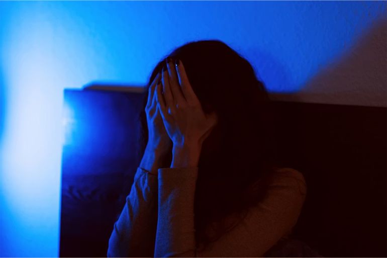 34-1784456605 A moody and evocative image of a person covering their face in distress, captured in low light with deep blue and orange tones. The photograph conveys a sense of emotional tension, isolation, or inner turmoil. Ideal for illustrating concepts related to mental health, anxiety, depression, loneliness, or difficult emotions in visual storytelling.