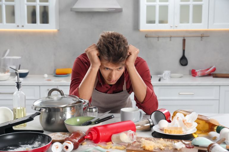 Upset man in messy kitchen. Many dirty dishware, utensils and food leftovers on table adobe stock