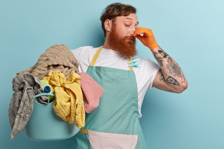 Foxy bearded man closes nose with fingers from unpleasant smell, collects all dirty laundry, wears casual t shirt and apron with clothespins, has tattoo, stands over blue wall, free space المصدر فري بيك