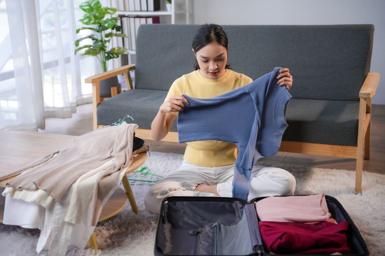 adobestock_869811350-1784648966 Woman happily packs clothes into a suitcase in her cozy, organized living room, preparing for an upcoming trip with excitement @ادوبي ستوك