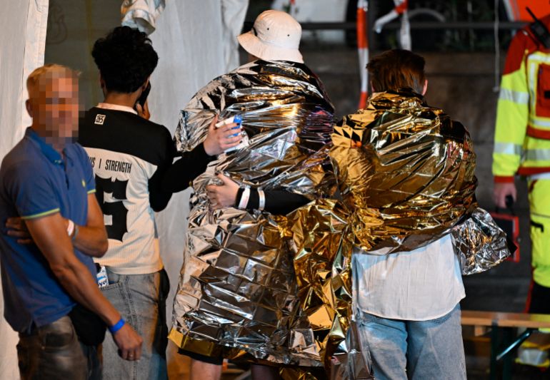 afp_6a6594fd0e0f-1785042173 People wrapped in security blankets hug near the scene where one person was killed and around 15 injured after a car is believed to have hit the crowd on the outskirts of the Christopher Street Day (CSD) parade in Berlin, on July 25, 2026.