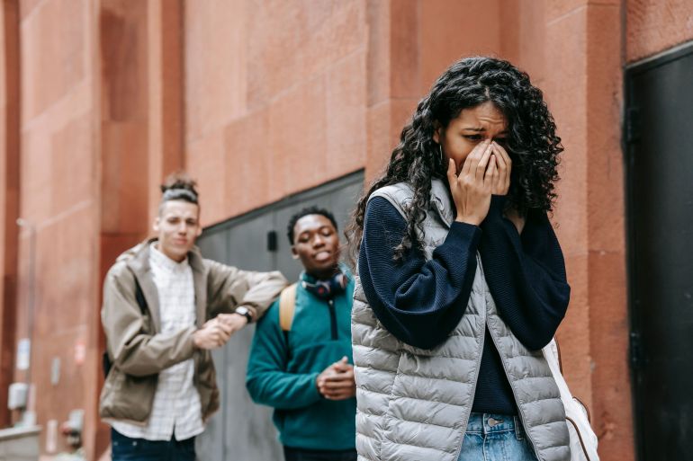 albkacabe-ghryb-1783201663 A teenager appears upset as two peers mock her in an urban setting, highlighting bullying issues.