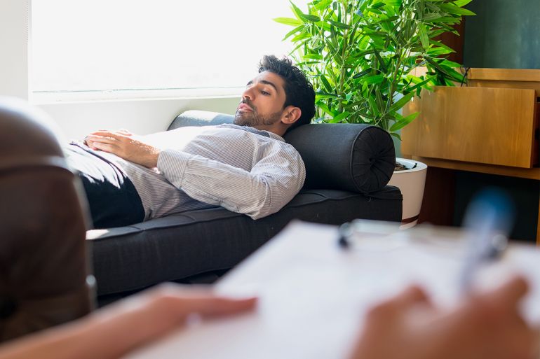 gettyimages-1320947589-1701707631 Portrait of a psychologist talking with his patient and taking notes while his lying on couch during a therapy session. Psychology and mental health concept.