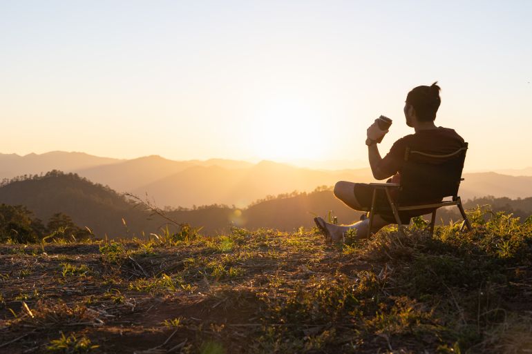 gettyimages-1370704812-1783028836 A man sitting on hill with cup of coffee. Morning and sunrise on beautiful nature. Mountains. happy man. inspiration concept, enjoy life. Calm and peace.