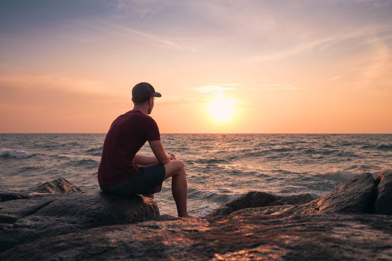 gettyimages-1430022513-1783028875 Rear view of man sitting on rock and watching idyllic sunset over ocean.