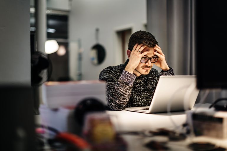 gettyimages-607477399-1784403878 Young casual businessman wearing glasses is sitting in front of his notebook holding his head pondering over his work. Office equipment and another computer is in front of him.