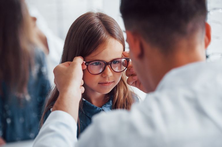 jhjh76-1783513937 Young pediatrician in white coat helps to get new glasses for little girl By standret @ادوببي ستوك