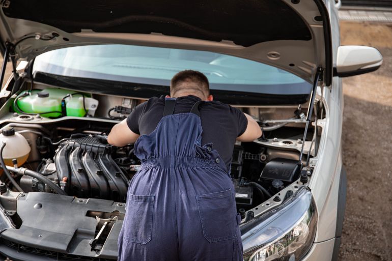 kk-1785162063 Mechanic in overalls working on a car engine outdoors, offering vehicle repair services.