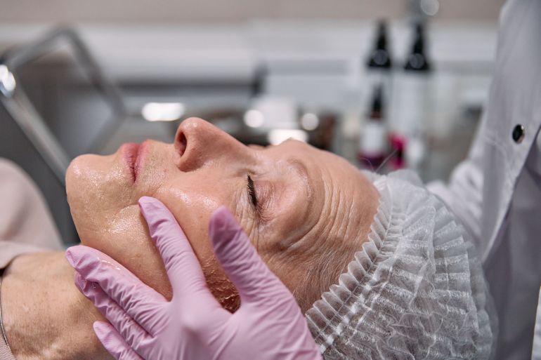 sh-1785081427 Beautician applies facial treatment on elderly woman with pink gloves in a clinic setting.