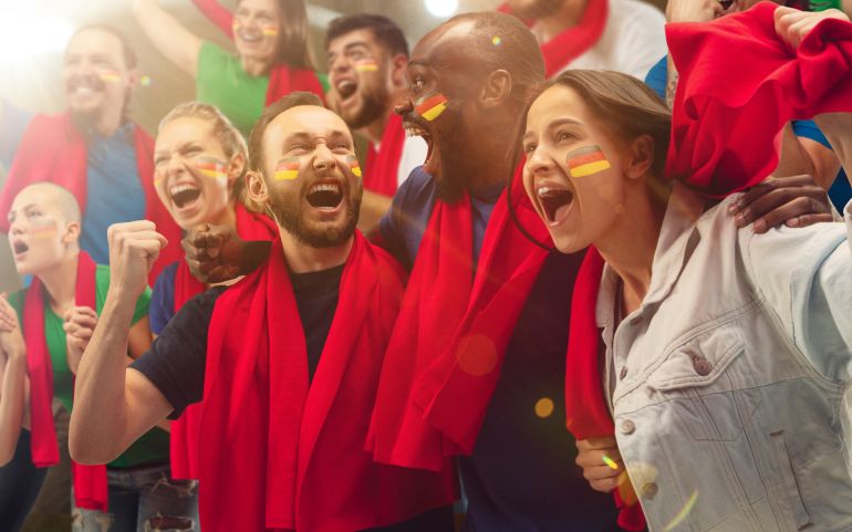 shutterstock_1919579174-1781101297 German football, soccer fans cheering their team with a red scarfs at stadium. Excited fans cheering a goal, supporting favourite players. Concept of sport, human emotions, entertainment.