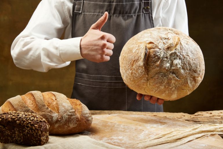 shutterstock_2340557791-1776254837 Male chef confectioner in apron hold warm homemade bread show thumb up studio background. Concept of food, health, traditions, bakery and ad