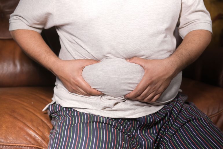 11911053-1786879847 A close-up view of a man holding his tummy while sitting on a couch, highlighting lifestyle choices.
