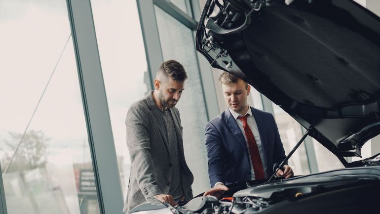 36729856-1786573312 Two men inspecting a car's engine hood in a dealership showroom.