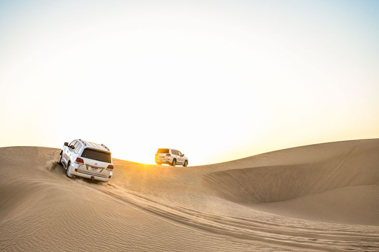 605-33923796-1786794286 White SUVs navigating the golden dunes at sunset in Sweihan, Abu Dhabi.