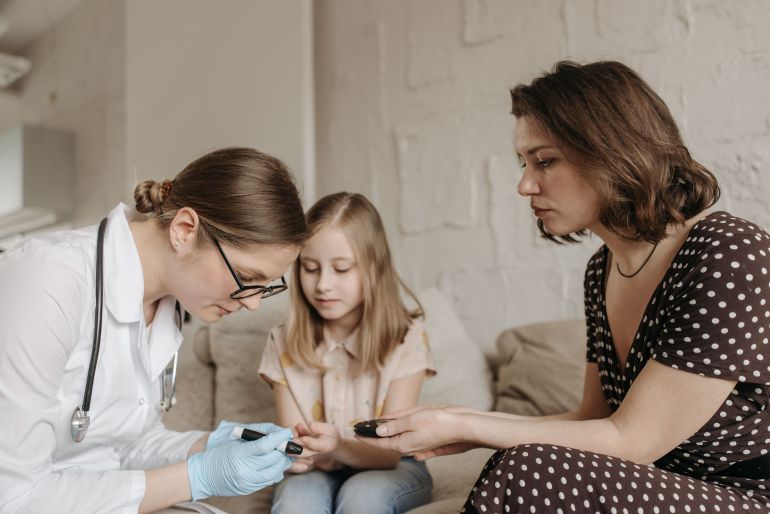 7653133-1787326356 A pediatrician checks a child's blood sugar levels while her mother assists at home.