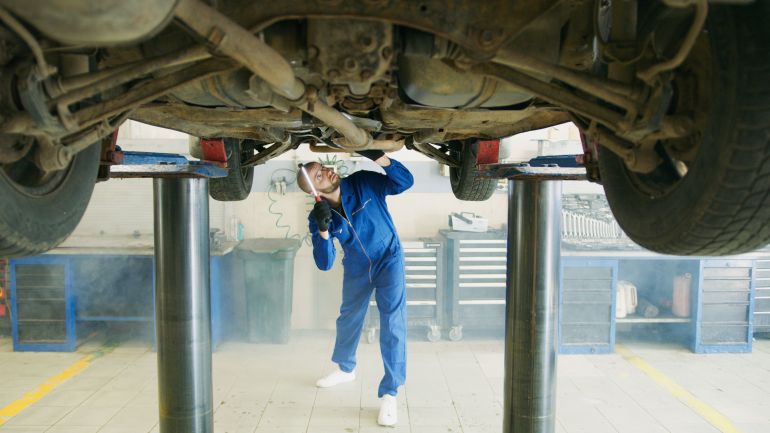 8985604-1786574423 A mechanic in blue uniform inspects a car lifted in a garage workshop.