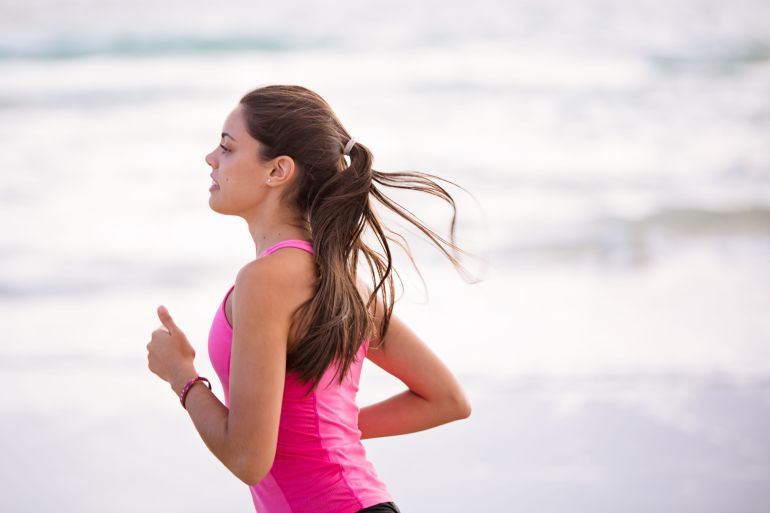 99590-1787228311 Side view of a young woman jogging on a beach wearing pink active wear.
