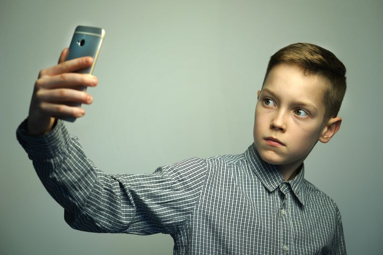 Teenage serious boy in office shirt with stylish haircut taking selfie on smartphone in studio.Adobe Stock
