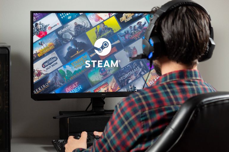 adobestock_17687942-1784535056 Close up to a male gamer at the right side of the picture holding a xbox controller watching a pc monitor with Steam video game platform @ادوبي ستوك