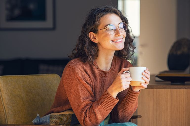adobestock_443783561-1787838458 Happy young woman drinking a cup of tea in an autumn morning. Dreaming girl sitting in living room with cup of hot coffee enjoying under blanket with closed eyes. Pretty woman wearing sweater at home and enjoy a ray of sunshine on a winter afternoon. المصدر: أدوبي ستوك