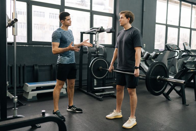 adobestock_813105135-1787402395 Wide shot of beginner sportsman doing barbell press during weightlifting workout with help of personal trainer at sport club. Young man exercising at gym support under supervision of coach. المصدر: أدوبي ستوك