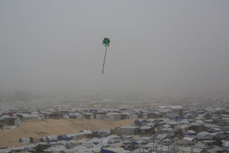 afp_6a8e11963379-1787695510 (FILES) A kite flies over tents and makeshift shelters at a camp for displaced Palestinians during hazy and dusty weather in Khan Yunis, in the southern Gaza Strip, on February 14, 2026.
